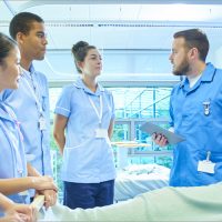 A  senior male staff nurse demonstrates the medical mannequin to a group of medical student nurses . They are all standing around the hospital bed listening to him .